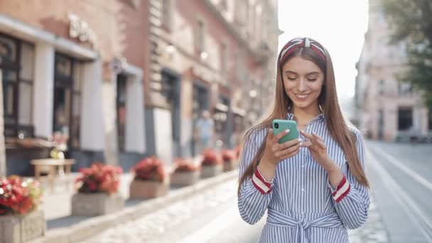파란색 과 흰색 줄무늬 옷을 입은 젊은 여성 이 스마트폰을 이용해 오래 된 거리를 걷고 있다. 소통, 소셜 네트워크, 온라인 쇼핑 개념. — 비디오