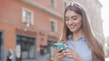Mavi ve beyaz çizgili gömlek giyen gülümseyen genç kadın eski sokak arka planda duran akıllı telefon kullanarak. İletişim, sosyal ağlar, online alışveriş konsepti.
