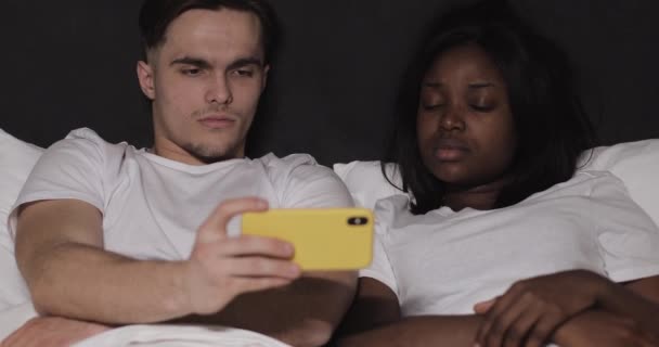 Pareja multiétnica viendo vídeo juntos en el teléfono inteligente amarillo acostado en la cama por la noche . — Vídeos de Stock