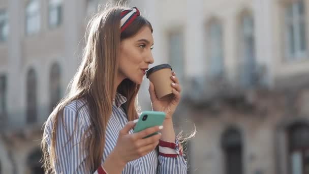 青と白のストライプのドレスシャツを着た美しい若い女性は、スマートフォンを使って古い通りに立ち、コーヒーを飲んでいます。コミュニケーション、ソーシャルネットワーク、オンラインショッピングの概念. — ストック動画