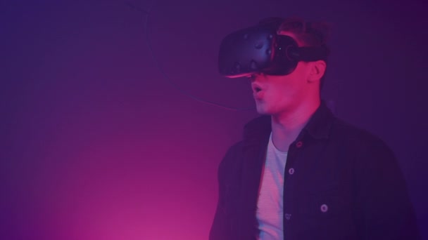 Jonge aantrekkelijke man met behulp van moderne Virtual Reality headset met gamepad op de achtergrond van neonlichten. Afstandsbedieningen, klikken, man werken in vr, te gebaren met de handen. — Stockvideo