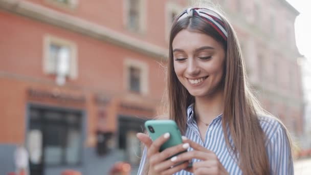 Χαριτωμένο γυναίκα φορώντας μπλε και λευκό ριγέ φόρεμα χρησιμοποιώντας το app για το smartphone στέκεται στην παλιά οδό της πόλης. Όμορφο κορίτσι που έχει καλά νέα στο smartphone. — Αρχείο Βίντεο