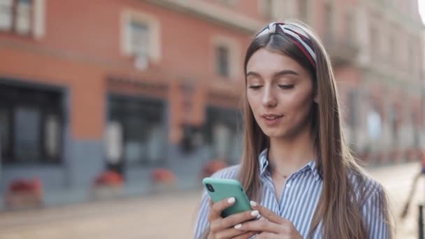 Χαριτωμένο γυναίκα φορώντας μπλε και λευκό ριγέ φόρεμα χρησιμοποιώντας το app για το smartphone περπάτημα στο παλιό δρόμο της πόλης. Όμορφο κορίτσι που έχει καλά νέα στο smartphone. — Αρχείο Βίντεο