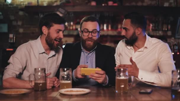 Три старых друга смотрят видео на смартфоне и пьют пиво в спортивном баре. Друзья смеются и обсуждают новости в пивном пабе . — стоковое видео