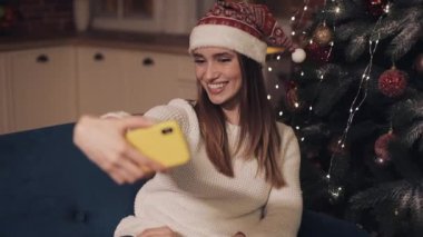 Happy Genç Çekici Kız Christamas Şapka Giyen Noel Ağacı yakınında Kanepede Oturan Tatil ve Yeni Yıl Selfie Poz Kavramı yapma.