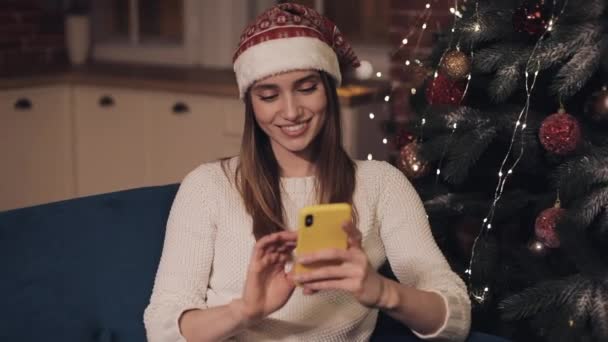 Bonita sorridente jovem senhora vestindo chapéu de Pai Natal sentado no sofá perto da árvore de Natal usando seu conceito de Smartphone de feriados e ano novo . — Vídeo de Stock