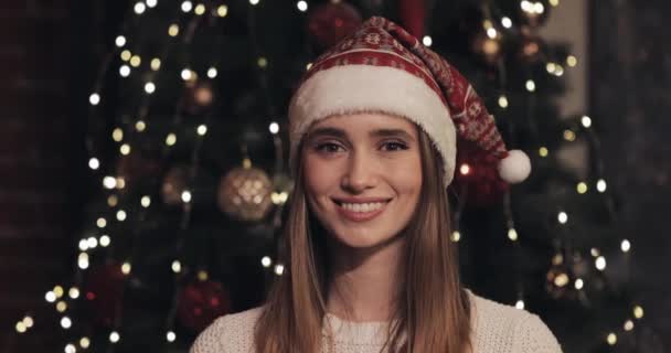 居心地の良いクリスマスの背景クローズアップでクリスマスツリーの近くに立ってサンタ帽子を身に着けている美しい若い白人女性の肖像画. — ストック動画