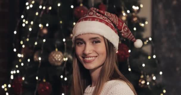 Retrato de hermosas sonrientes mujeres caucásicas jóvenes con sombrero de Santas de pie cerca del árbol de Navidad en el acogedor fondo de Navidad y girando de cabeza a la cámara de cerca . — Vídeos de Stock