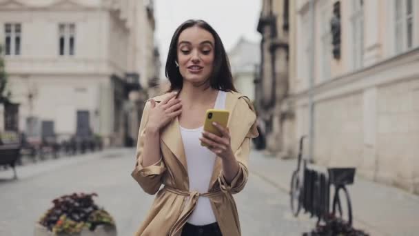 Preety gelukkig jong meisje dragen draadloze koptelefoon het maken van een video gesprek glimlachend praten wandelen ontspannen op de straat stad achtergrond close-up. — Stockvideo
