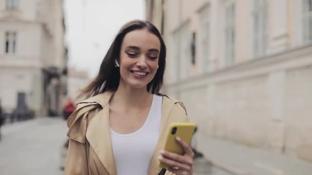 Menina bonita feliz usando fones de ouvido sem fio fazendo uma chamada de vídeo Smiling Talking acenando com a mão andando Relaxado no fundo da cidade de rua Close Up . — Vídeo de Stock