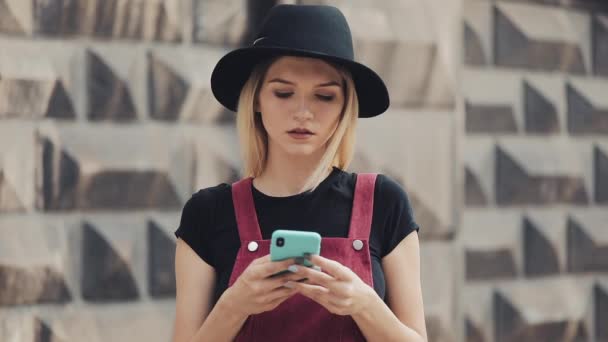 Portret van jong blond meisje met piercing in de neus het dragen van een zwarte hoed met behulp van haar Smartphone staande op de stad straat close-up. — Stockvideo