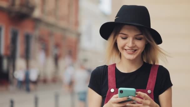 Jovem Preety Loira com piercing no nariz vestindo um chapéu preto usando seu smartphone andando sorrindo na rua da cidade  . — Vídeo de Stock