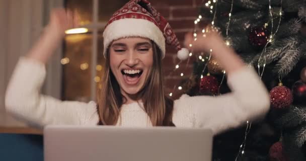 Jóvenes hermosas mujeres con sombrero de Navidad sentadas en el sofá en el fondo del hogar usando su computadora portátil se regocija sonríe y ríe aplaude su concepto de las manos de las vacaciones y el año nuevo — Vídeo de stock