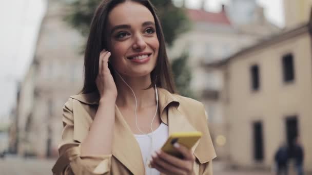 Hermosa chica joven que usa auriculares disfruta escuchando música usando su teléfono inteligente y sonriendo caminando en el fondo de la ciudad . — Vídeo de stock