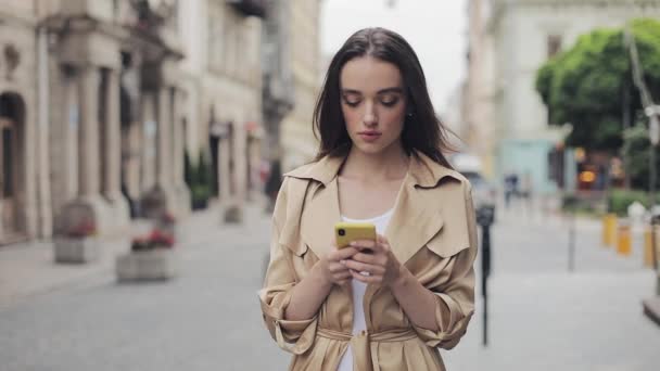 Portret van mooie jonge meisje houden mobiele telefoon chatten staande loking in de camera en glimlachend op de achtergrond van de stad. — Stockvideo