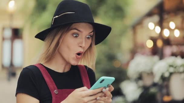 Jovem bonita menina loira com anel de nariz vestindo um chapéu preto usando seu smartphone surpreso dizendo Wow e Gesturing sim de pé na rua da cidade . — Vídeo de Stock