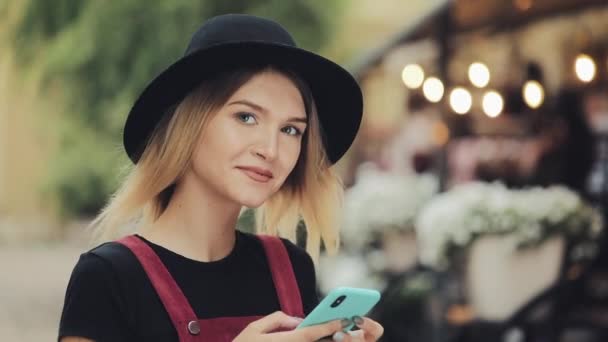 Retrato de una joven rubia bonita con un anillo en la nariz con un sombrero negro usando su teléfono inteligente parado en la calle City sonriendo a la cámara de cerca Zoom  . — Vídeos de Stock