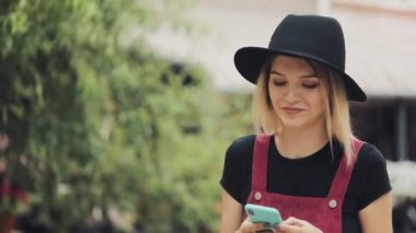 Genç İyi Görünümlü Gülümseyen Sarışın Kız Siyah Şapka Giyen Onu Smartphone Typing ve City Arka Planda Yürüyüş kullanarak.