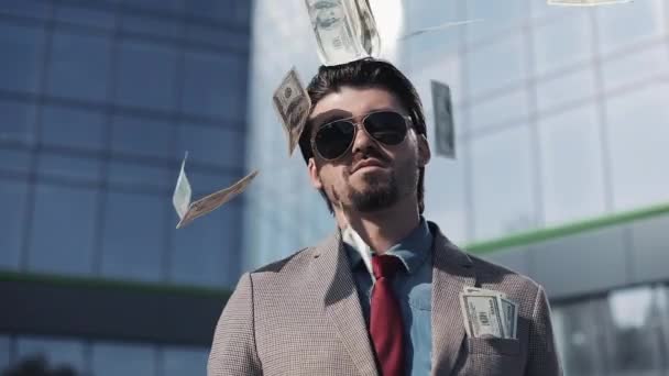 Homme riche en costume moderne portant des lunettes de soleil debout dans la rue près de l'immeuble de bureaux tandis que des tas d'argent tombent de près — Video