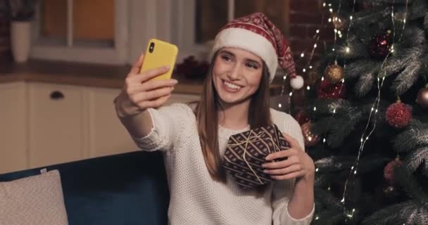 Hermosa mujer con sombrero de Christamas sentada en el sofá sosteniendo y sacudiendo la caja de regalo cerca del árbol de Navidad haciendo una videollamada o mensaje de vídeo Concepto de días festivos y año nuevo de cerca — Vídeo de stock