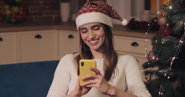 Güzel Genç Sarışın Kafkas Kız Santas Şapka Giyen Noel Ağacı yakınında oturan onu Smartphone ve Tatil ve Yeni Yıl Close Up Gülümseyen Kavramı kullanarak. — Stok video