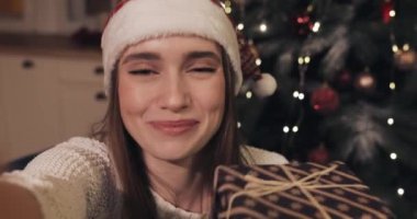 Güzel Kadın Giyen Christamas Şapka Evde Noel Ağacı yakınında Oturan Holding ve Sallayarak Hediye Kutusu Tebrik Tatil ve Yeni Yıl Bir Video Arama veya Video Mesaj Kavramı yapma.