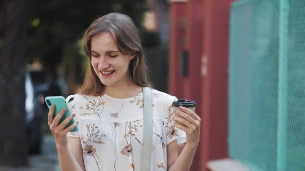 Schöne junge kaukasische braune Haare Mädchen trägt weißes Blumenkleid mit Tasche über der Schulter mit ihrem Smartphone Kaffee trinken in der Nähe von türkisfarbenem Zaun lächelnd. — Stockvideo