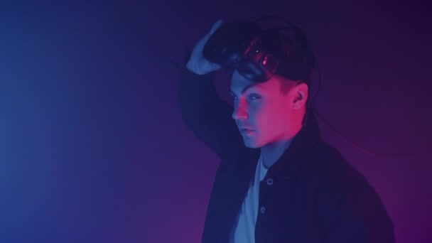 Porträt eines jungen attraktiven Mannes, der sein Virtual-Reality-Headset abnimmt und in die Kamera blickt. Hintergrund der Cyberbeleuchtung. — Stockvideo