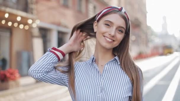 Jeune fille heureuse et souriante aux cheveux bruns et au bandeau élégant portant une robe rayée échangeant les cheveux et souriant regardant la caméra debout à la rue de la ville Zoom sur . — Video