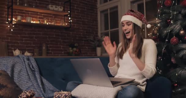 Sorrindo atraente jovem caucasiano menina vestindo chapéu Santas sentado perto da árvore de Natal em casa fundo usando seu laptop conversando texting digitação alegra sorrisos e gargalhadas aplausos suas mãos — Vídeo de Stock