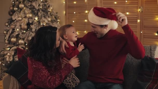 居心地の良いホームホリデーの背景でクリスマスツリーの近くのソファに座ってママのお父さんと愛らしい男の子の陽気な家族。お父さんは休日と新年の彼の帽子の概念を使用して息子と遊びます. — ストック動画