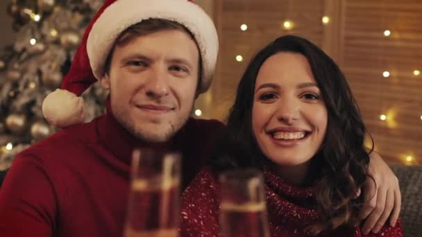居心地の良い家の背景の笑顔でクリスマスツリーの近くのソファに座っている幸せな若いカップルの肖像画は、休日と新年のカメラの概念にシャンパンのメガネを上げるクローズアップ. — ストック動画