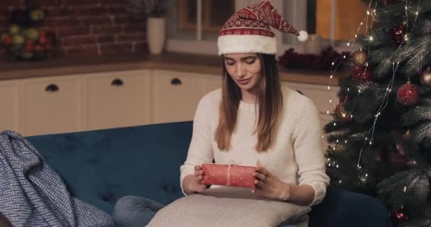 Молоді жінки в капелюсі Сантас сидять на дивані на затишному Різдвяному тлі тримаючи червоний подарунок Радісна концепція свят та Нового року крупним планом . — стокове відео