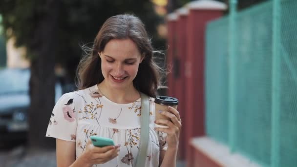 Šťastné usmívající se mladá preety dívka s hnědými vlasy chůze po ulici používání jejího mobilního telefonu s kávovou šálkou, která vypadá spokojeně a spokojeně. — Stock video