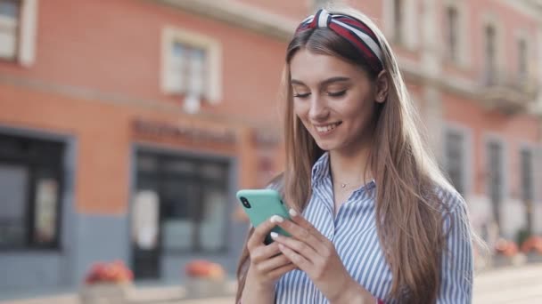 Unga snygga attraktiva kaukasiska flicka med brunt hår och pannband på den med hennes smartphone rullning och leende stående på City Street. — Stockvideo