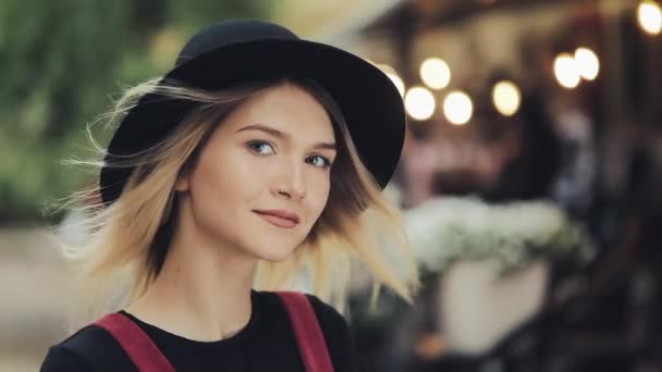 Portrait de jeune jolie fille blonde avec anneau nasal portant un chapeau noir vent soufflant ses cheveux debout à la rue de la ville sourire à la caméra Gros plan Zoom  . — Video
