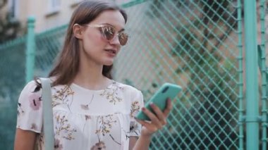 Genç Pretty Kahverengi Saç Kafkas Kız Onun Smartphone kullanarak Güneşli Gün Dışında Yürüyüş Şık Güneş Gözlüğü Giyen Close Up. İletişim kavramı.