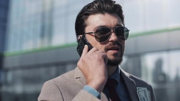 Junger gutaussehender Geschäftsmann, der Anzug und Sonnenbrille trägt und zufrieden am Telefon spricht und Geldscheine in der Nähe des Geschäfts aus nächster Nähe hält — Stockvideo