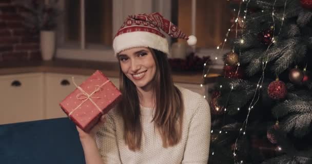 Portret van jonge mooie vrouwen dragen Santas hoed zittend op een bank in de buurt van de kerstboom op gezellige kerst achtergrond houden en schudden van een rode huidige doos en lachende zoom. — Stockvideo