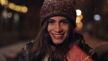 Kış Şapka ve Eldivenler Genç Charming Happy Girl Close Up Portre, Holding ve Sarılma Present Box, Falling Snow Ayakta, Evening Street Lights Arka Planda Kameraya Gülümseyen.