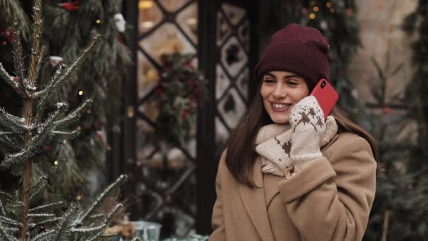 Sorpreso bella ragazza in inverno cappello e guanti, parlando su smartphone, dicendo Wow, ridendo mentre in piedi all'aperto a Natale decorato sfondo. Comunicazione, Concetto Vacanze . — Video Stock