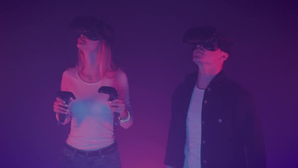 Молоді пари носять віртуальну реальність гарнітура, дівчина Холдинг джойстики контролери, стоячи озираючись в кімнаті з прохолодною кольору неонових. — стокове відео