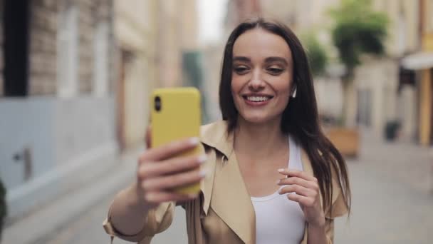 イヤホンを身に着けている魅力的な若い女性は、ビデオ通話を行い、電話を垂直に話し、街の通りの背景を歩いて笑うクローズアップ. — ストック動画