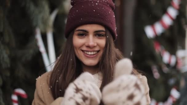 Retrato da jovem menina bonita feliz em roupas de inverno em pé em flocos de neve caindo Fora, sorrindo, dançando no Natal decorado janela loja fundo. Conceito de férias de inverno . — Vídeo de Stock