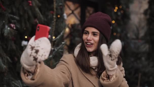 Joven hermosa chica sonriente con sombrero de invierno, bufanda, manoplas, hacer videollamada o mensaje de vídeo, ondeando, hablando, de pie al aire libre en el fondo decorado de Navidad. Concepto de vacaciones de invierno . — Vídeo de stock