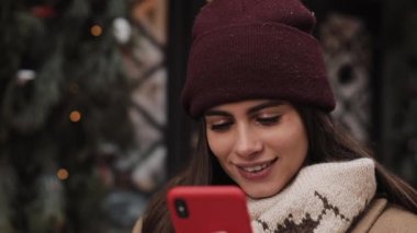 Kışın Gülen Pretty Girl Close Up Hat ve Mittens Sohbet, Onun Smartphone kullanarak, Ekran a looking, Açık Ayakta. İletişim, Noel ve Yılbaşı Tatili Konsepti.