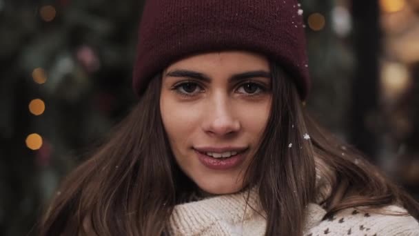 Close up Ritratto di giovane bella ragazza caucasica che indossa cappello invernale e sciarpa, In piedi in caduta fiocchi di neve fuori, Sorridente, Guardando alla macchina fotografica. Concetto vacanza invernale . — Video Stock