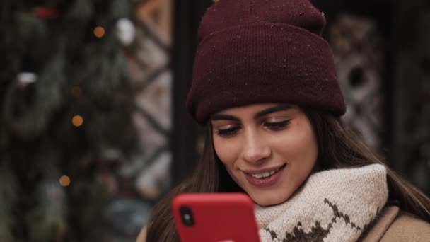 Nahaufnahme eines lächelnden hübschen Mädchens mit Wintermütze und Fäustlingen, die mit ihrem Smartphone chatten, auf den Bildschirm schauen und draußen stehen. Kommunikation, Weihnachts- und Neujahrsferien-Konzept. — Stockvideo