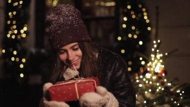 Kış Şapka ve Mittens Içinde Gülümseyen Preety Girl Kırmızı Present Box Holding, Mutlu Görünümlü, Arka Planda Işıklar ve Christmas City Sokak Süslemeleri Ayakta. Mutlu Tatil Konsepti.