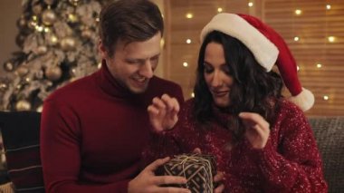 Yakışıklı Koca ve Onun Çekici Karısı Bir Hediye Kutusu üzerinde Bir Yay Bağlı bitirdi ve Tatil ve Yeni Yıl Evde Arka Plan Kavramı Noel Ağacı yakınında Kanepede Oturan Öptü.